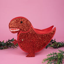 Load image into Gallery viewer, The kawaii red glitter dino vegan bag on a pink studio background with green foliage. The vegan leather red sparkly bag is facing forward to highlight the red glittered side, dinosaur face, gold metal detailing, detachable strap and moveable arm.
