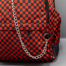 Load image into Gallery viewer, Close up of the Red Checkerboard Backpack sat on a grey background. The vegan friendly bag is facing forward to highlight the red and black check print, two front zip pockets, two elasticated side pockets, main top double zip pocket and silver draping decorative chain.
