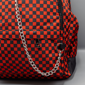 Close up of the Red Checkerboard Backpack sat on a grey background. The vegan friendly bag is facing forward to highlight the red and black check print, two front zip pockets, two elasticated side pockets, main top double zip pocket and silver draping decorative chain.