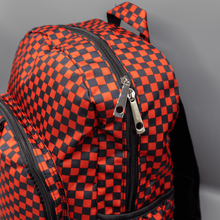 Load image into Gallery viewer, Close up of the Red Checkerboard Backpack sat on a grey background. The vegan friendly bag is facing forward to highlight the red and black check print, two front zip pockets, two elasticated side pockets, main top double zip pocket and silver draping decorative chain.
