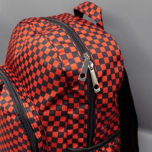 Close up of the Red Checkerboard Backpack sat on a grey background. The vegan friendly bag is facing forward to highlight the red and black check print, two front zip pockets, two elasticated side pockets, main top double zip pocket and silver draping decorative chain.
