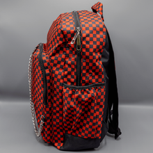 Load image into Gallery viewer, The Red Checkerboard Backpack sat on a grey background. The vegan friendly bag is facing left to highlight the red and black check print, two front zip pockets, two elasticated side pockets, main top double zip pocket and silver draping decorative chain.
