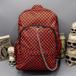 The Red Checkerboard Backpack sat on a grey background with a palmistry guide book and two skull stack on the right and a phrenology guide candle and three skull stack on the left. The vegan friendly bag is facing forward to highlight the red and black check print, two front zip pockets, two elasticated side pockets, main top double zip pocket and silver draping decorative chain.