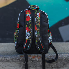 Load image into Gallery viewer, Skulls and roses vegan backpack sat on a skatepark bench in front of a graffiti wall. The bag is facing away to highlight the skulls and roses printed padded adjustable shoulder straps, two elasticated side pockets and plain black back.
