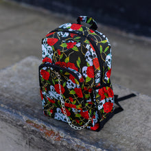 Load image into Gallery viewer, The skulls and roses vegan backpack sat on a skatepark bench. The bag is facing forward angled to the left to highlight the skulls and roses all over print, silver chain, two zip front pockets and two side elastic pockets.
