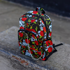 The skulls and roses vegan backpack sat on a skatepark bench. The bag is facing forward angled to the left to highlight the skulls and roses all over print, silver chain, two zip front pockets and two side elastic pockets.