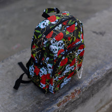 Load image into Gallery viewer, The skulls and roses vegan backpack sat on a skatepark bench. The bag is facing forward angled to the right to highlight the skulls and roses all over print, silver chain, two zip front pockets and two side elastic pockets.
