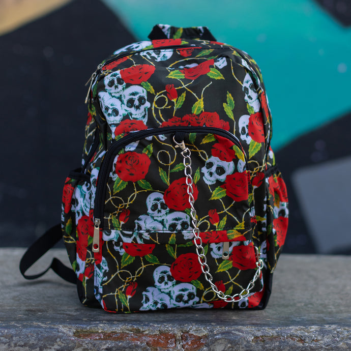 Skulls and roses vegan backpack sat on a skatepark bench in front of a graffiti wall. The bag is facing forward to highlight the skulls and roses all over print, silver chain, two zip front pockets and two side elastic pockets.