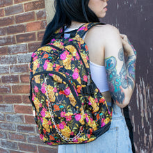 Load image into Gallery viewer, Model standing in front of a brick wall wearing the floral gold skull nylon chain vegan backpack. The bag is facing the camera to highlight the flowers and skulls print, two zipped pockets, two elastic side pockets and detachable decorative silver chain.
