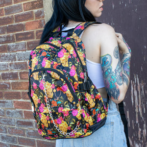 Model standing in front of a brick wall wearing the floral gold skull nylon chain vegan backpack. The bag is facing the camera to highlight the flowers and skulls print, two zipped pockets, two elastic side pockets and detachable decorative silver chain.