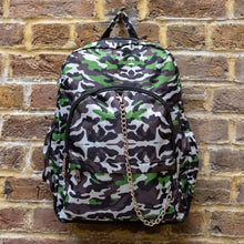 Load image into Gallery viewer, The Savannah Camouflage Backpack hanging outside on a brick wall. The green, brown, khaki, cream and black vegan friendly backpack is facing forward to highlight the two front zip pockets with a silver draping detachable chain, the two side elasticated pockets, the top handle and the main double zip compartment.
