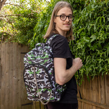 Load image into Gallery viewer, Jack is stood in a garden area modelling the savannah camouflage camo vegan backpack. The bag is facing towards the camera to highlight the front camo print, two front zip pockets, two elastic side pockets, detachable silver chain and top handle.
