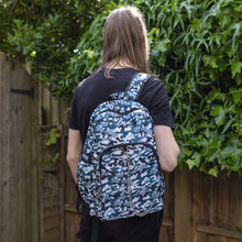 Load image into Gallery viewer, Jack is stood in a garden area modelling the snow camouflage camo vegan backpack. The bag is facing towards the camera to highlight the front camo print, two front zip pockets, two elastic side pockets, detachable silver chain and top handle.
