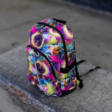 Load image into Gallery viewer, The pink and purple space galaxy vegan backpack sat on a skatepark bench. The backpack is facing forward angled left to highlight the multicoloured space/planet/galaxy print, two zip pockets, two elastic side pockets and detachable silver chain.
