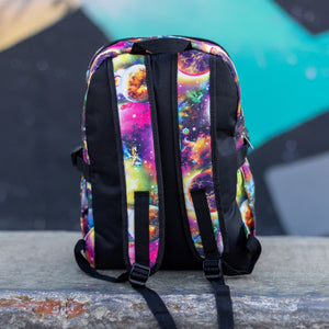 The pink and purple space galaxy vegan backpack sat on a skatepark bench. The backpack is facing away from the camera to highlight the two side pockets, the plain black back and two padded adjustable shoulder straps.