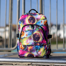 Load image into Gallery viewer, The pink and purple space galaxy vegan backpack sat at the top of a skatepark ramp. The backpack is facing forward to highlight the multicoloured space/planet/galaxy print, two zip pockets, two elastic side pockets and detachable silver chain. 
