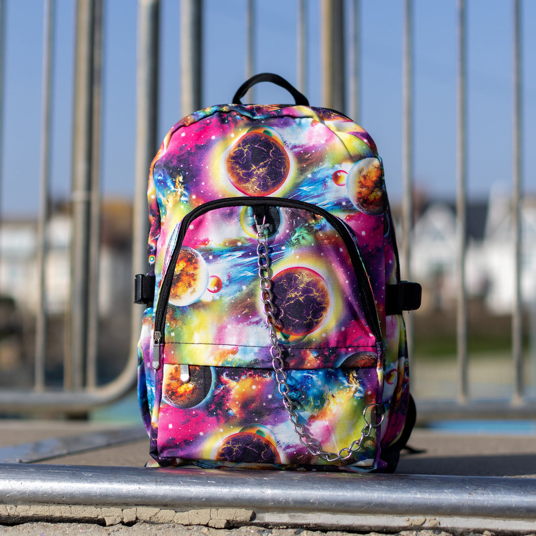 The pink and purple space galaxy vegan backpack sat at the top of a skatepark ramp. The backpack is facing forward to highlight the multicoloured space/planet/galaxy print, two zip pockets, two elastic side pockets and detachable silver chain. 