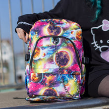 Load image into Gallery viewer, The pink and purple space galaxy vegan backpack sat at the top of a skatepark ramp next to a model. The backpack is facing forward to highlight the multicoloured space/planet/galaxy print, two zip pockets, two elastic side pockets and detachable silver chain. 
