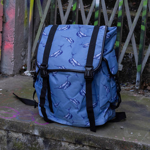 The Blue Swordfish Vegan backpack sat near a graffiti urban area. A pastel blue backpack with black buckle and strap detailing with swordfish printed all over. The bag is facing forward to highlight the side buckle pockets, drawstring and buckle close and black piping detailing.