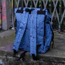 Load image into Gallery viewer, The Blue Swordfish Vegan backpack sat near a graffiti urban area. A pastel blue backpack with black buckle and strap detailing with swordfish printed all over. The bag is facing away from the camera to highlight the side zip pockets, side buckle pockets and adjustable shoulder straps.

