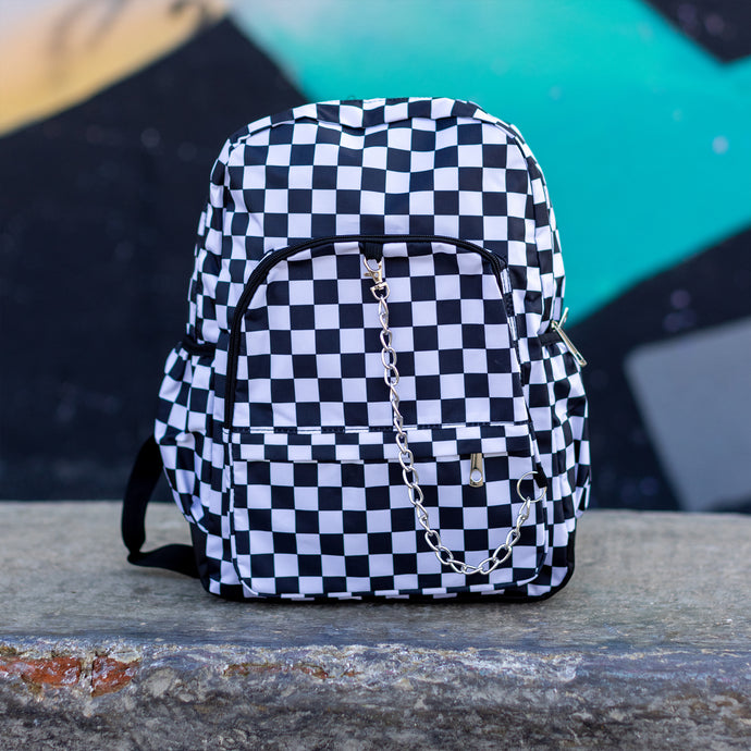 CHOK white checker vegan backpack sat on a skatepark bench in front of a graffiti wall. The bag is facing forward to highlight the white and black checkered square print, silver detachable chain, two front zip pickets and two side elastic pockets.