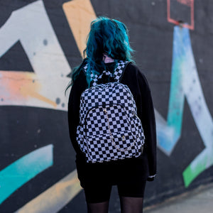 Model with blue hair wearing all black is stood in a skatepark near a graffiti wall wearing the chok white checker vegan backpack.