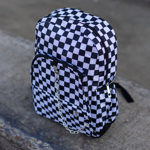 CHOK white checker vegan backpack sat on a skatepark bench. The bag is facing forward angled left to highlight the white and black checkered square print, silver detachable chain, two front zip pickets and two side elastic pockets.