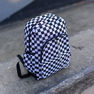 CHOK white checker vegan backpack sat on a skatepark bench. The bag is facing forward angled right to highlight the white and black checkered square print, silver detachable chain, two front zip pickets and two side elastic pockets.