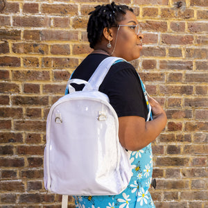 The White & Clear Window Ita Backpack being worn on a shoulder of a model wearing run and fly dungarees and a black tshirt. The bag is facing forward to highlight the clear front window with two metal D rings, two side elasticated pockets, main zip compartment and top handle. The vegan friendly bag is inspired by kawaii jfashion.
