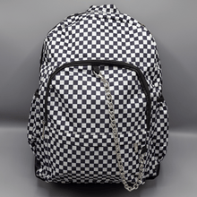 Load image into Gallery viewer, The White Checkerboard Backpack sat on a grey background. The vegan friendly bag features an all over mini black and white check print with black trim along the silver zips. The bag is facing forward to highlight the two front zip pockets, two side elasticated pockets, the top zip compartment, the top handle and the silver decorative detachable chain draping across the front.
