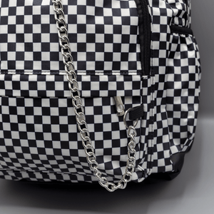 Close up of the White Checkerboard Backpack sat on a grey background. The vegan friendly bag features an all over mini black and white check print with black trim along the silver zips. The bag is facing forward to highlight the two front zip pockets, two side elasticated pockets, the top zip compartment, the top handle and the silver decorative detachable chain draping across the front.