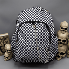 Load image into Gallery viewer, The White Checkerboard Backpack sat on a grey background with a palmistry guide book and two stacked skulls on the left and a phrenology candle and three stacked skulls on the right. The vegan friendly bag features an all over mini black and white check print with black trim along the silver zips. The bag is facing forward to highlight the two front zip pockets, two side elasticated pockets, the top zip compartment, the top handle and the silver decorative detachable chain draping across the front.
