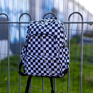 The chok white checker vegan backpack is hanging on a skatepark railing. The bag is facing forward to highlight the white and black checkered square print, silver detachable chain, two front zip pickets and two side elastic pockets.