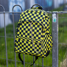 Load image into Gallery viewer, The chok yellow checker vegan backpack is hanging on a skatepark railing. The bag is facing forward to highlight the yellow and black checkered square print, silver detachable chain, two front zip pickets and two side elastic pockets.
