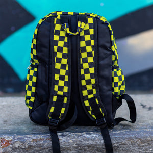 CHOK yellow checker vegan backpack sat on a skatepark bench in front of a graffiti wall. The bag is facing away to highlight the yellow and black checkered square printed and adjustable padded shoulder straps, two elastic side pockets and plain back.