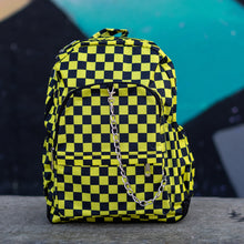 Load image into Gallery viewer, CHOK yellow checker vegan backpack sat on a skatepark bench in front of a graffiti wall. The bag is facing forward to highlight the yellow and black checkered square print, silver detachable chain, two front zip pickets and two side elastic pockets.
