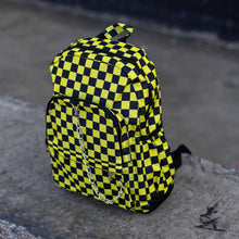 Load image into Gallery viewer, CHOK yellow checker vegan backpack sat on a skatepark bench. The bag is facing forward angled left to highlight the yellow and black checkered square print, silver detachable chain, two front zip pickets and two side elastic pockets.
