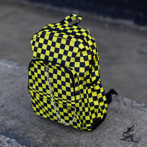 CHOK yellow checker vegan backpack sat on a skatepark bench. The bag is facing forward angled left to highlight the yellow and black checkered square print, silver detachable chain, two front zip pickets and two side elastic pockets.