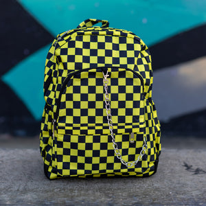 CHOK yellow checker vegan backpack sat on a skatepark bench in front of a graffiti wall. The bag is facing forward to highlight the yellow and black checkered square print, silver detachable chain, two front zip pickets and two side elastic pockets.