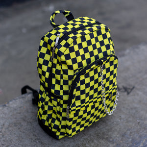 CHOK yellow checker vegan backpack sat on a skatepark bench. The bag is facing forward angled right to highlight the yellow and black checkered square print, silver detachable chain, two front zip pickets and two side elastic pockets.