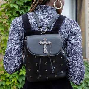Yolande, an alternative pierced model, wearing a spiderweb long sleeve top with a black pinafore and the GothX Don't Cross Me Tassel Tie Backpack. They are wearing the vegan leather bag on their back with both detachable straps to highlight the cross, skull and hexagonal studs, tassel tie close and the cross chain emblem magnetic clip close flap.